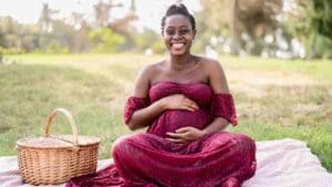 Mulher negra retinta grávida de vestido vinho, sentada em um lençol na grama, com uma cesta de piquinique, segurando a barriga e sorrindo em direção a câmera